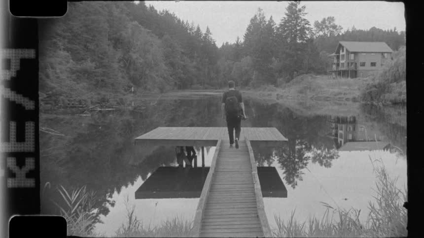 Man with a backpack and holding a camera walks on a wooden deck at a lake
