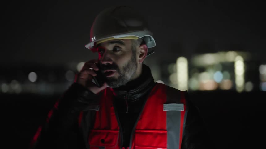 Engineer wearing a hard hat and reflective vest answers a call at night on a construction site
