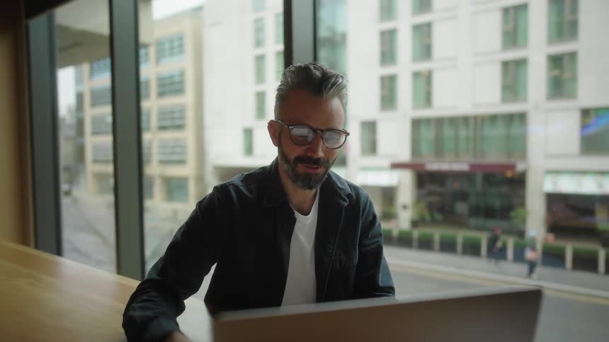 Man in glasses waves at laptop screen during video call