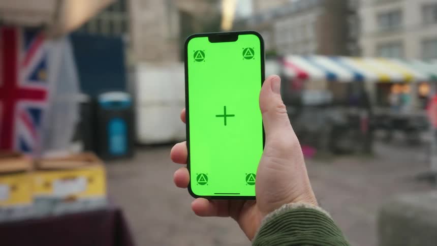 A hand is holding a smartphone with a bright green screen displaying a crosshair and four triangular symbols in front of a colorful street market