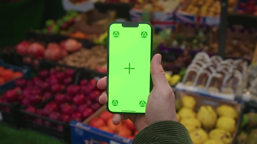 A hand is holding a smartphone with a bright green screen displaying a crosshair and four triangular symbols in front of a colorful fruit and vegetable market stall