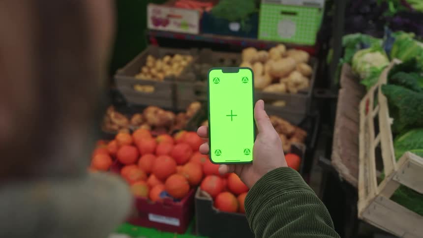 Person holds a smartphone in front of boxes of fruits and vegetables at an openair market