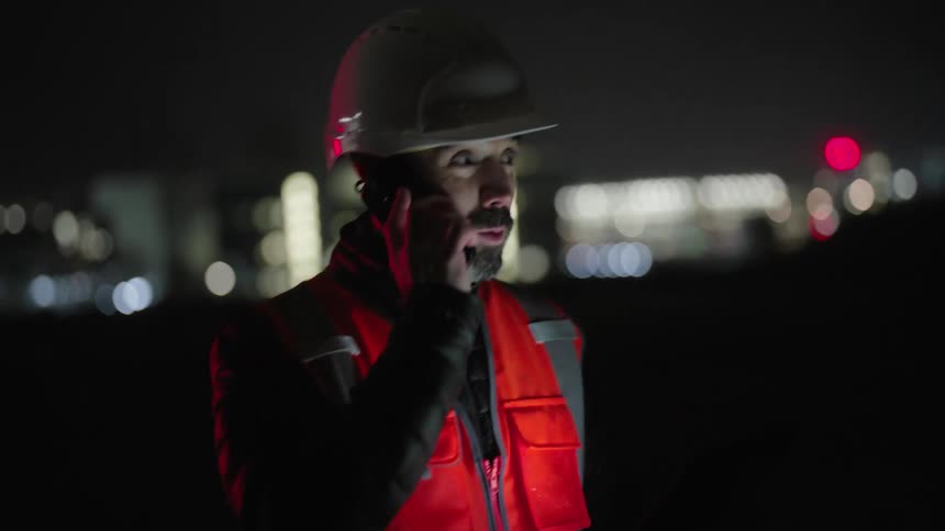 Man in helmet and protective vest is on phone and talking outdoors at night