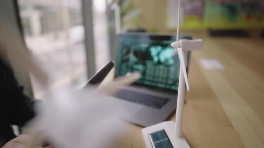 A person inspects a small model of a wind turbine while using a tablet next to a laptop on a desk by a window with a view of the street outside