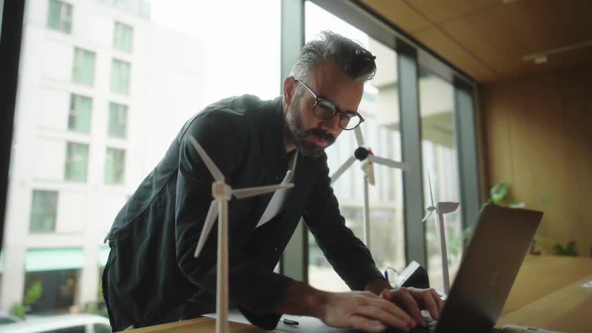 A man is standing using a laptop and then takes off his glasses
