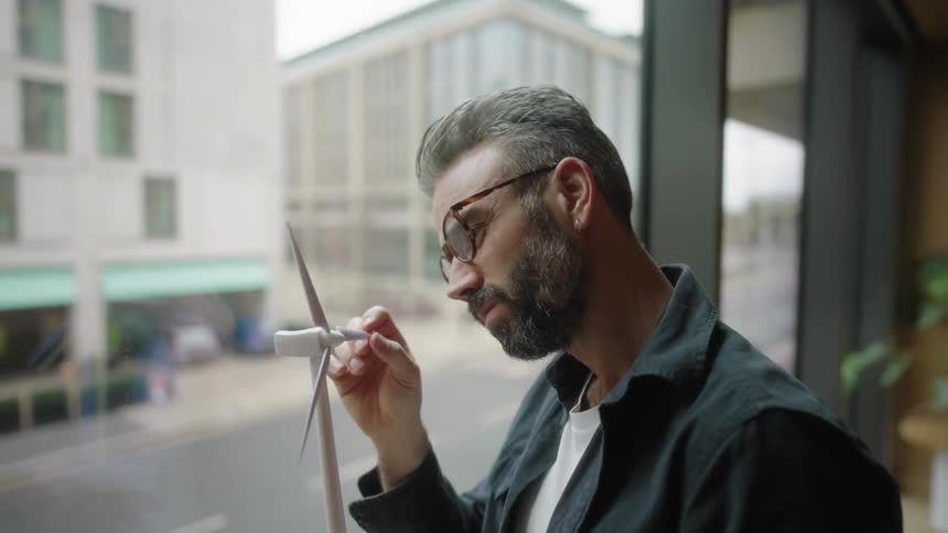 A person is holding and inspecting at a small model of a wind turbine while sitting by a window with a view of a street outside