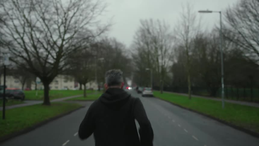A person is jogging down the middle of a treelined street with cars parked along the sides and houses visible in the background