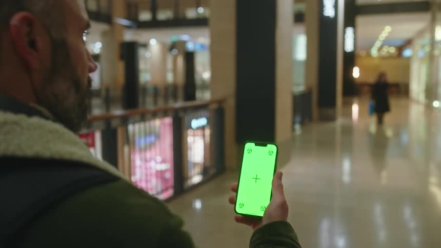 A person is holding a smartphone with a bright green screen displaying a plus sign in the center while standing in a welllit modern indoor shopping mall