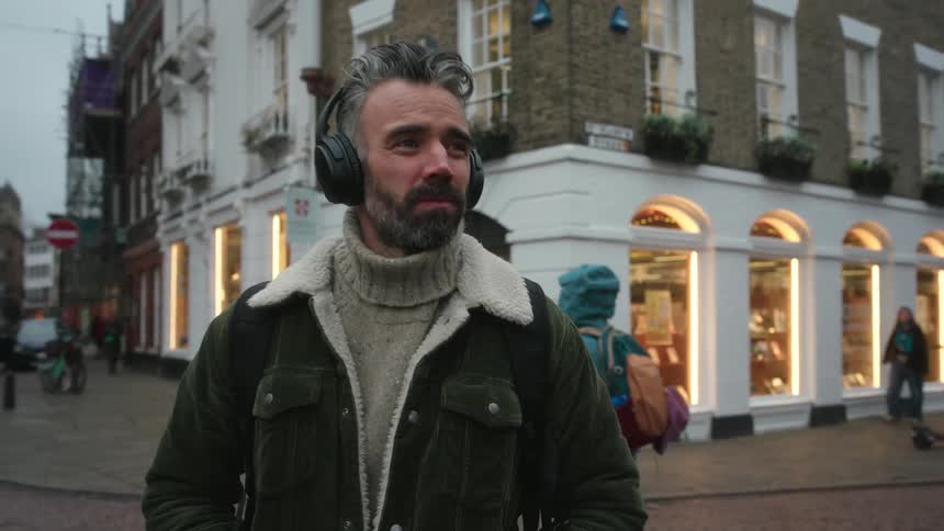 Man in a green coat and black suitcase watches the movement on a cobblestone street and puts on headphones