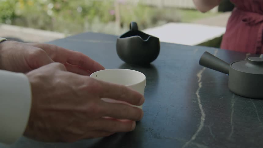 Two people interact around a white cup on a dark table with a teapot in the background