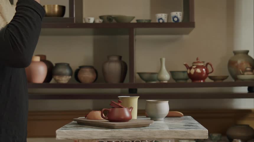 Person in longsleeved shirt holds a small cup on a marble table with a teapot and sweets in a room decorated with ceramics