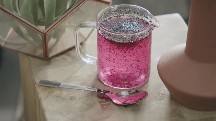 A clear glass pitcher filled with a pink liquid and dark specks sits on a table next to a spoon with a similar pink substance