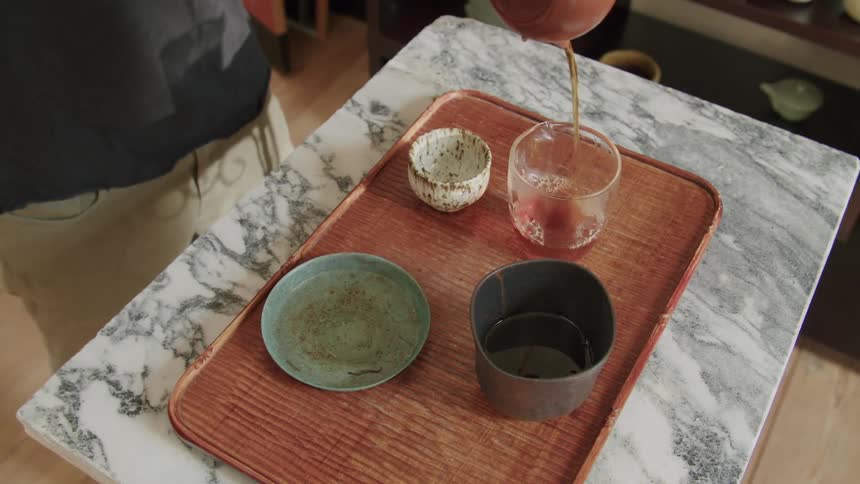Person pours tea from a brown teapot into a glass cup on a wooden tray on a marble table