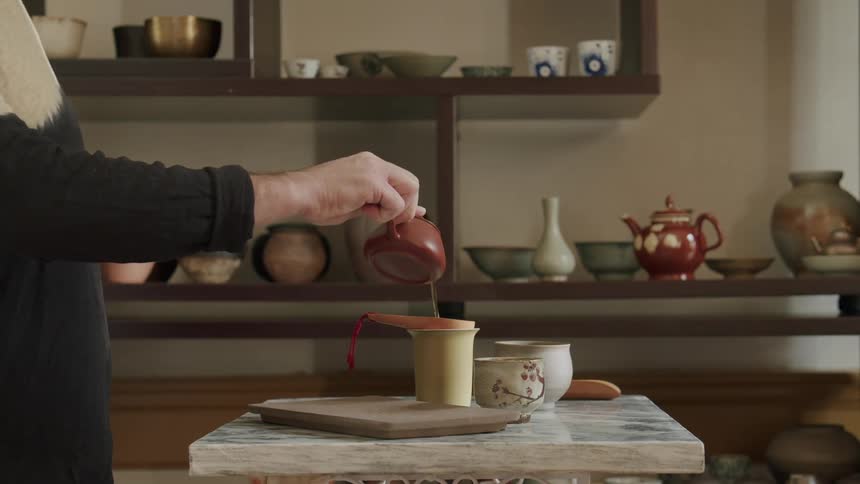 Person puts tea leaves in a small ceramic teapot then pours tea from a brown teapot into a filtered cup on the table