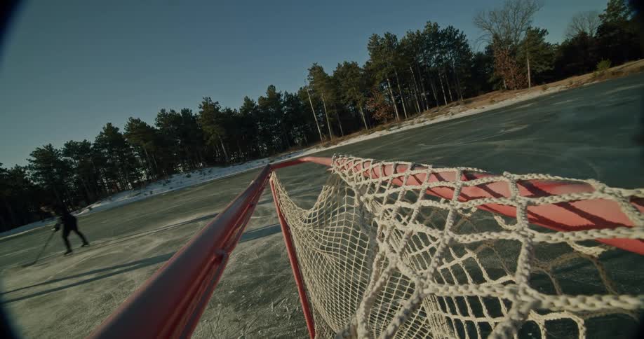Two hockey players dressed in jackets beanies and gloves and carrying hockey sticks skate away on a frozen lake after adjusting the hockey goal net with trees surrounding the icy rink