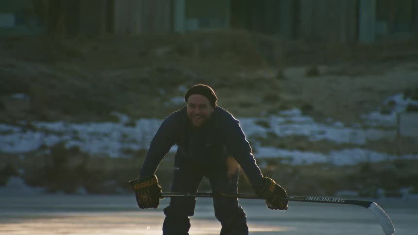Man in jacket beanie pajamas skates and gloves squats on frozen lake anticipating a shot with hockey stick as steam rises from his breath in the chilly dawn