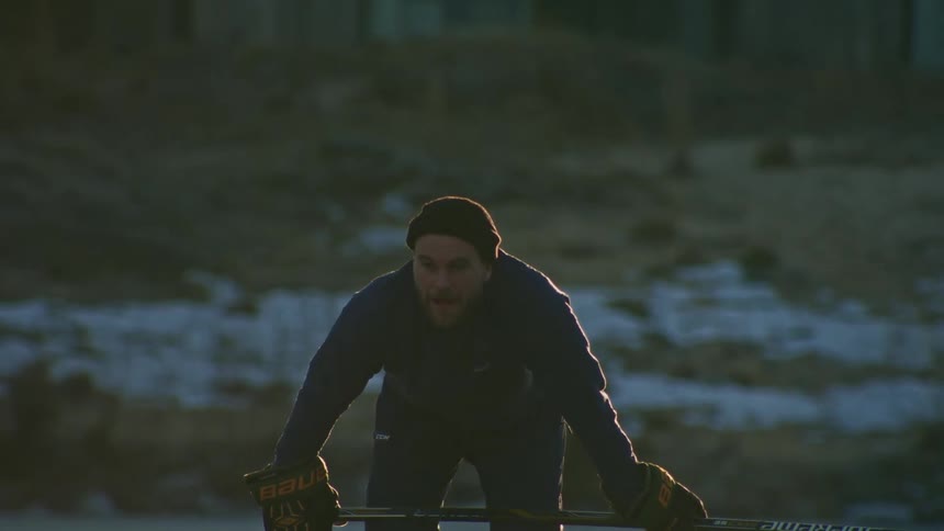 On a frozen lake a man stands hunched over breath steaming in the cold holding a hockey stick with gloved hands signifying a chilly winter day