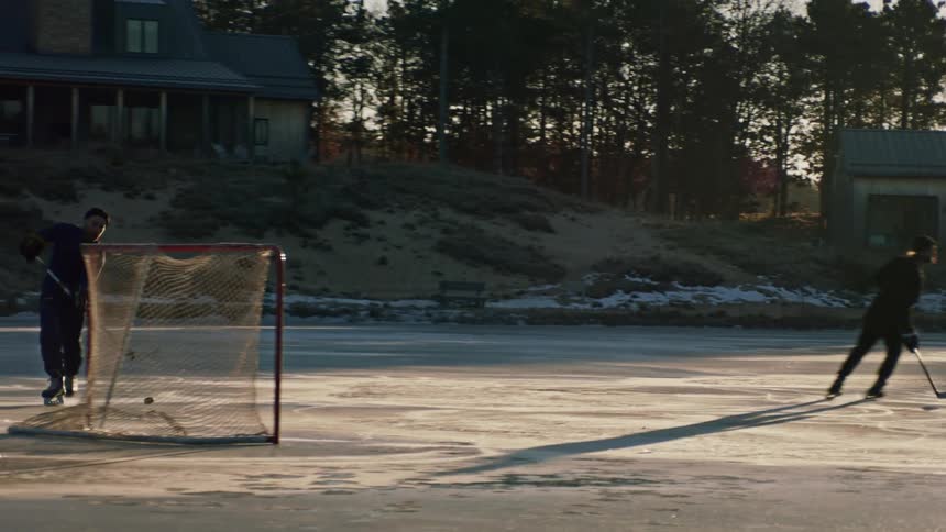 Two people are playing ice hockey on an outdoor rink with a house and trees visible in the background during a sunny winter day