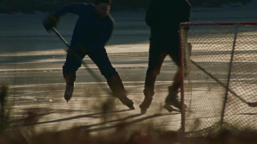 Two people play ice hockey on a frozen outdoor surface at dusk one skates forward controlling the puck with a stick while the other stands near the goal silhouetted against the reflective ice and a blurred snowy background