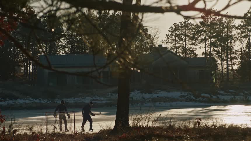 Two people playing hockey on an outdoor ice rink with a house in the background