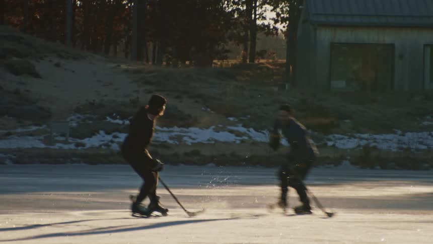 Two men in winter clothing play ice hockey on a frozen lake outdoors with one attempting to score on the other