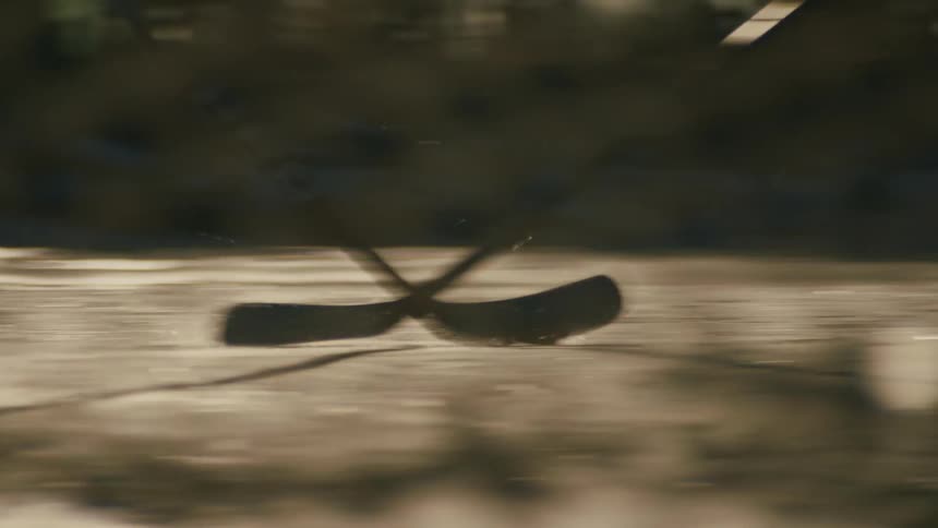 Hockey players skate rapidly and pass a puck on an outdoor rink with fencing and a red post briefly visible