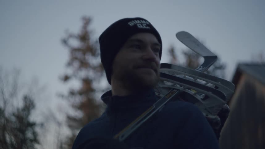 A man wearing a beanie and carrying hockey skates and a stick over his shoulder smiles in an outdoor setting with trees and a building in the background