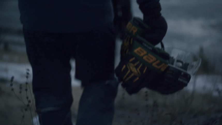 Two people walk towards a frozen lake one carrying a hockey helmet and gloves in a wintery setting