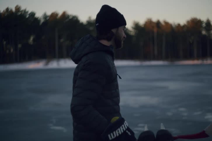 A man walking near a frozen lake in the morning carrying hockey sticks and skates