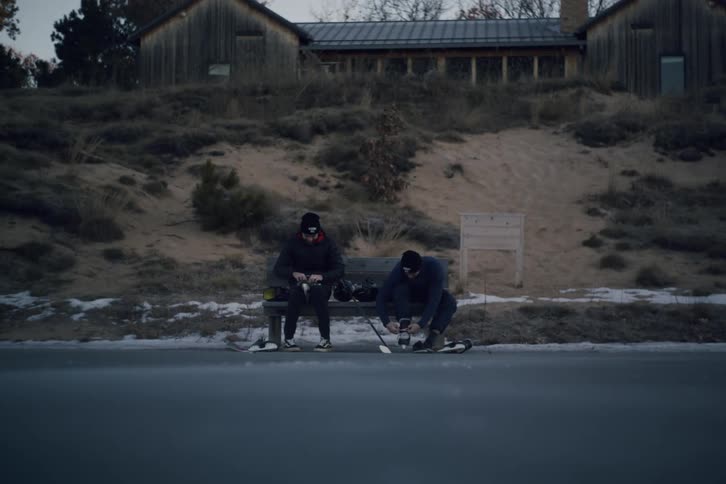 Two friends sitting on bench outdoors on a frozen pond lacing up hockey skates