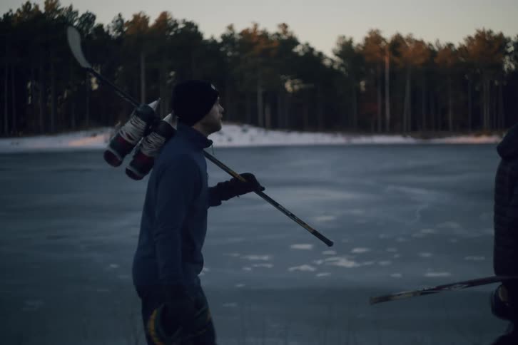 Two people walking near a frozen lake in the morning carrying hockey sticks and skates over their shoulders