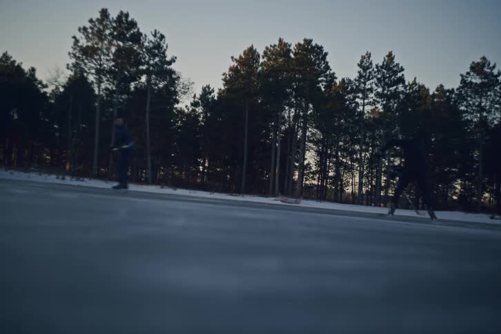 Two hockey players skate on an outdoor rink as the sun sets behind a line of tall pine trees