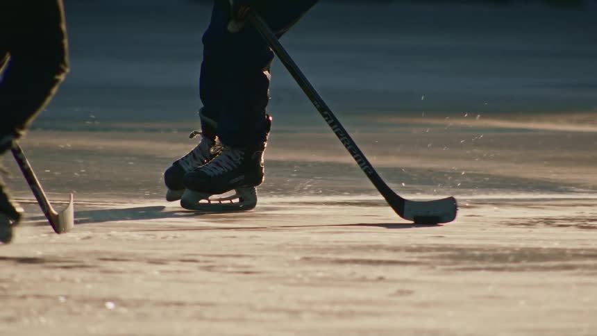 Two people shown from waist to feet play ice hockey on frozen water