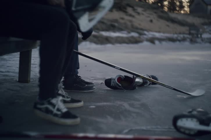 Two people sit on a bench and prepare to put on hockey skates with gloves and sticks lying nearby