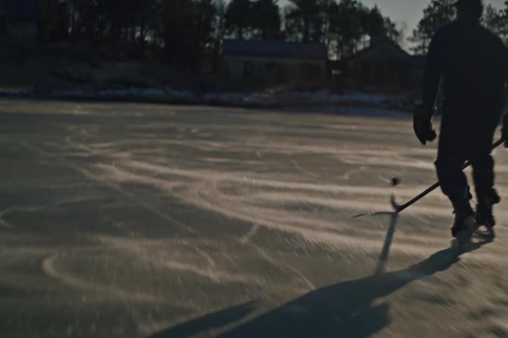 A person silhouetted against a dimly lit ice rink playing hockey and casting long shadows across the ice