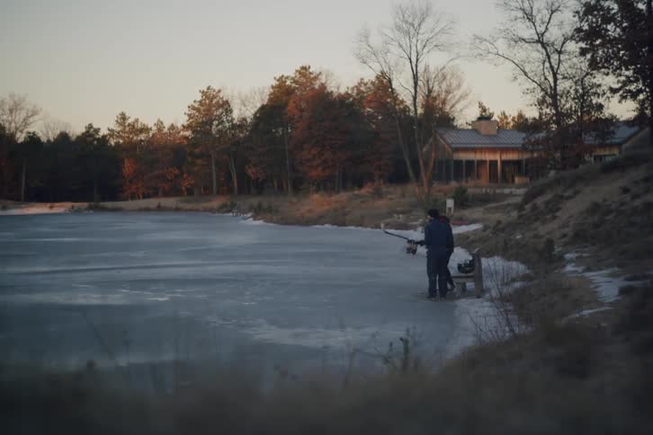 A person carrying hockey gear walks along the snowy bank of a frozen lake towards a bench with a house and trees in the background