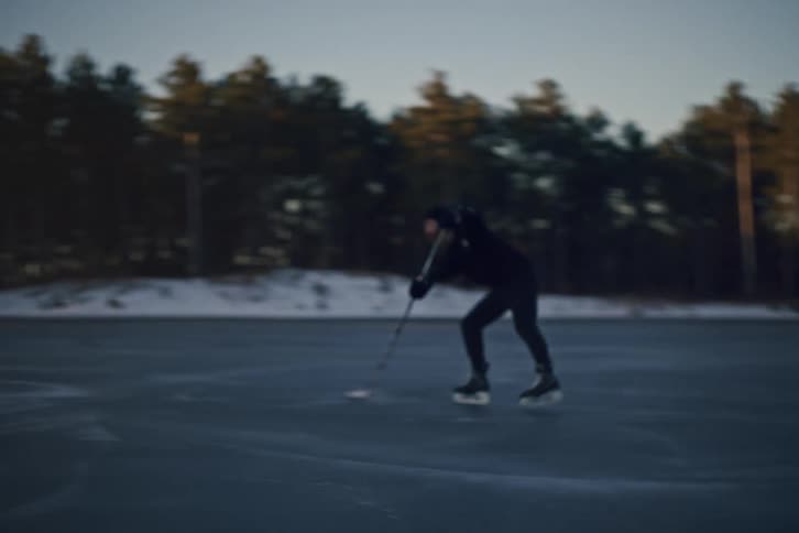 A hockey player skates and stickhandles a puck on an outdoor rink with trees in the background