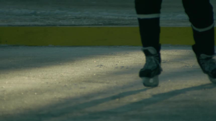 Several ice hockey players skating on an outdoor rink focusing on their lower bodies and equipment one player in a red jersey handles a hockey stick while others in dark clothing move closely with the ice reflecting the fading light