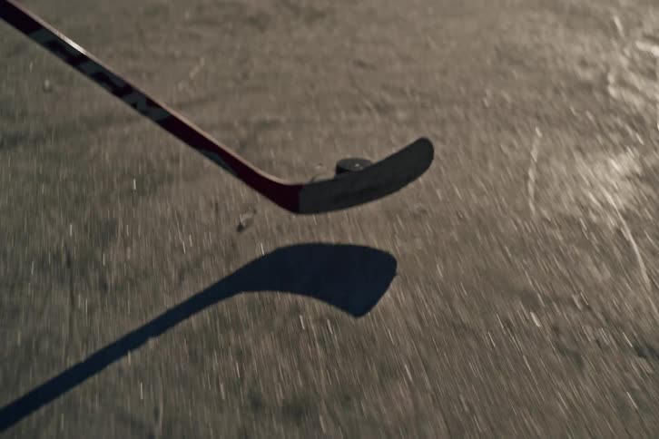 A hockey stick is being driven toward a puck on a scratched ice surface