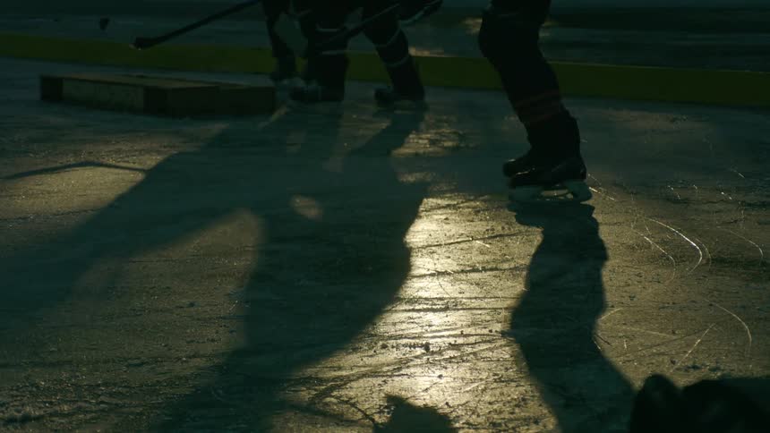 People silhouetted against a dimly lit ice rink playing hockey and casting long shadows across the ice