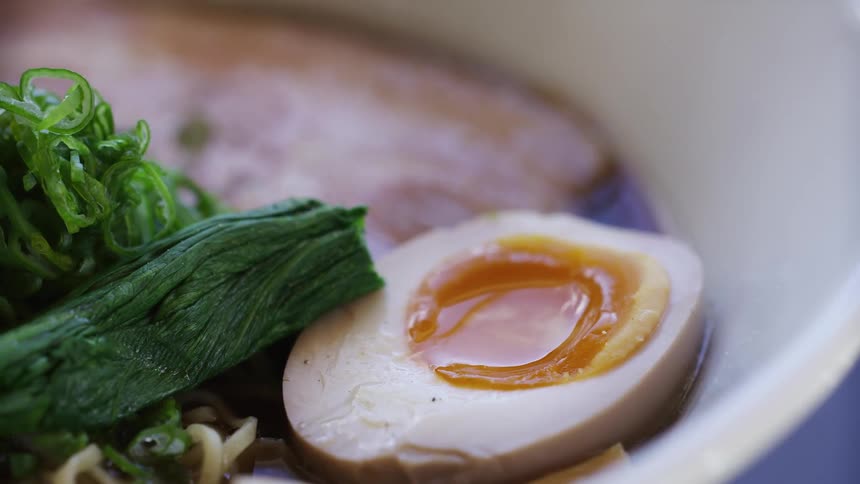 A closeup shot shows a half of a softboiled egg with a bright orange yolk next to chopped green onions and a dark green vegetable in a bowl of ramen