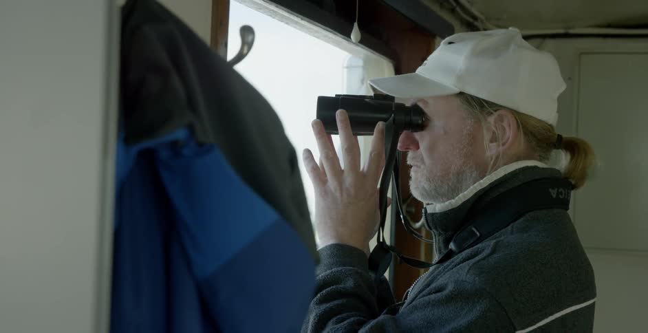 Bearded man looking out a window through his binoculars