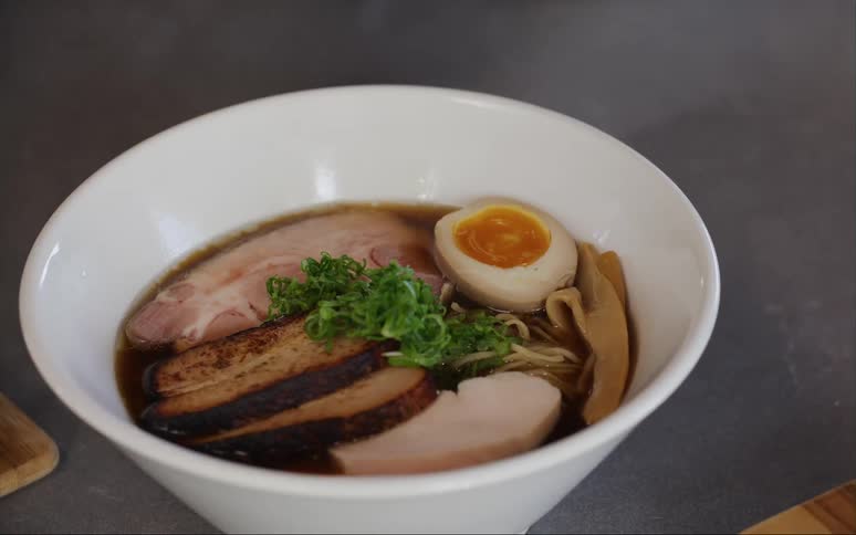 Hand finishes a bowl of ramen by placing a boiled egg and seaweed on top of the dark broth