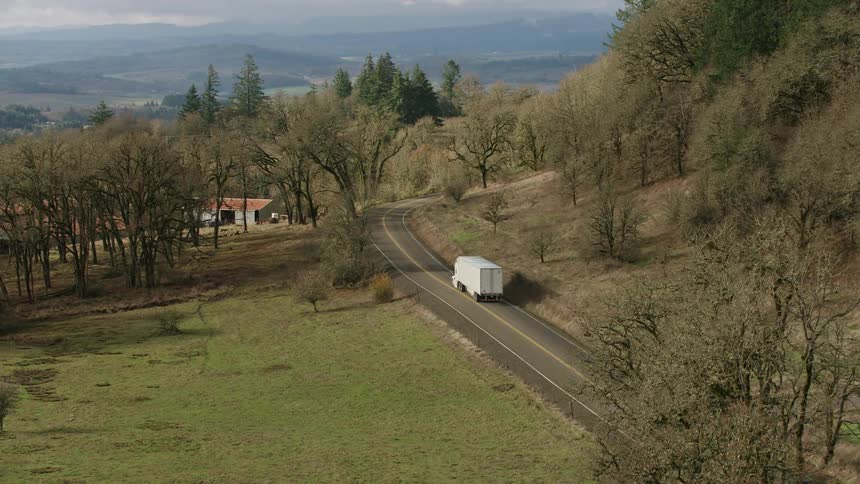Trucks travel along winding country roads amid hills and trees under a cloudy sky