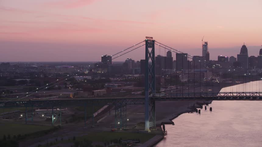 Illuminated suspension bridge stretches over a river at dusk with the city in the background