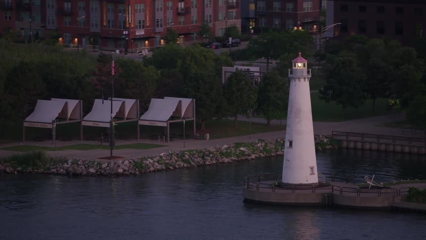 An aerial shot shows a white lighthouse with a lit lamp standing on a breakwater in a harbor at dusk