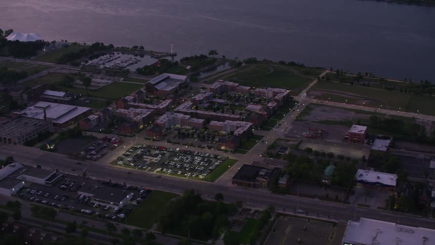 An aerial view shows a city neighborhood at dusk with buildings streets and a brightly lit parking lot visible