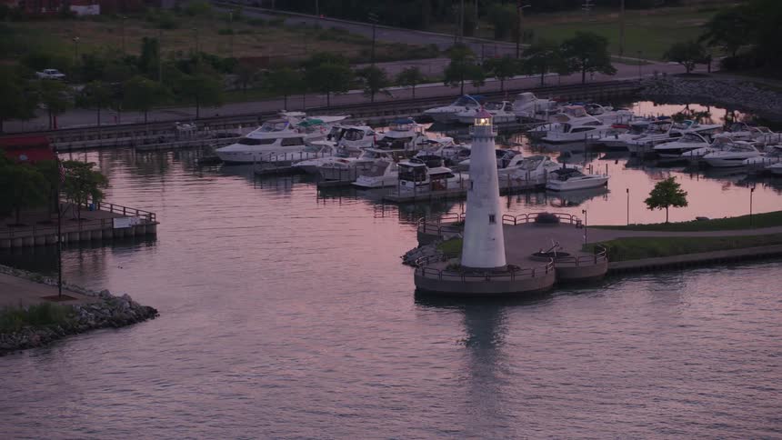 An aerial shot shows a white lighthouse with a lit lamp standing on a breakwater in a harbor at dusk