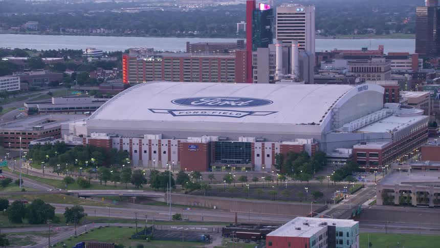 An aerial view shows ford field stadium in detroit with a river and cityscape in the background