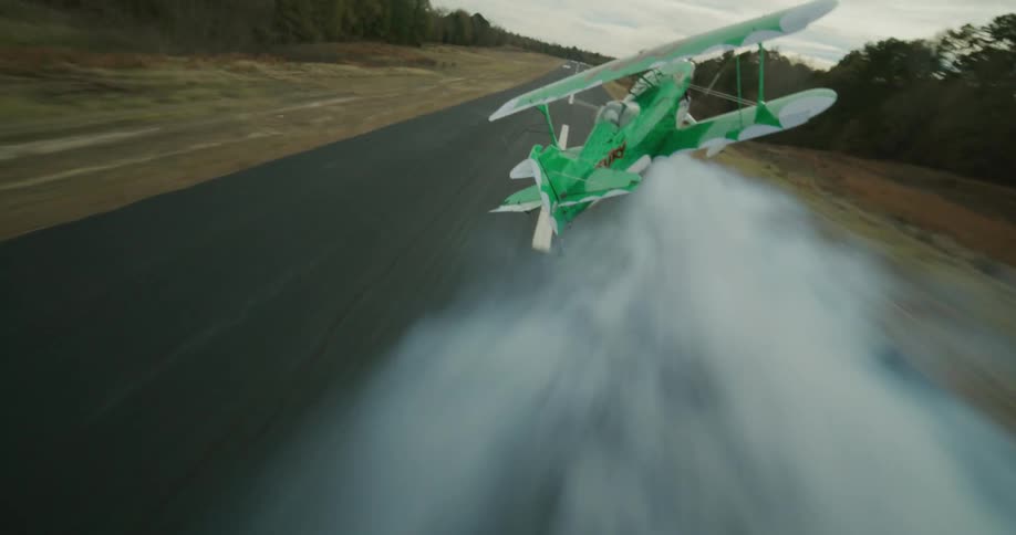 Green biplane with the word fury painted on the upper wing emits smoke while it takes off from a runway bordered by the woods